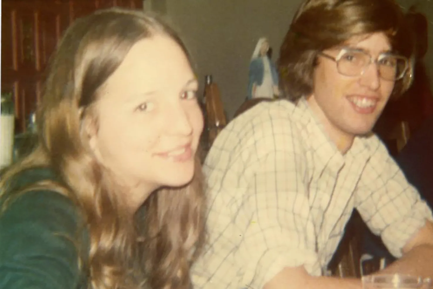 In this 1970s photo, a young white couple smiles at the person taking the picture, almost as if they're saying "if you really have to take this photo, go ahead." He wears glasses and she has long hair--their hair color almost matches.