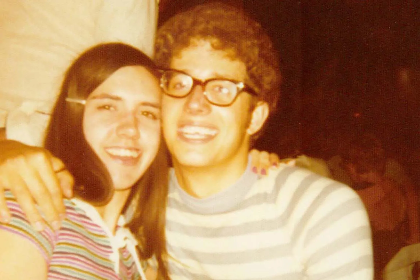 The same couple from the same era grin as they pose for a photo. They both wear striped shirts.