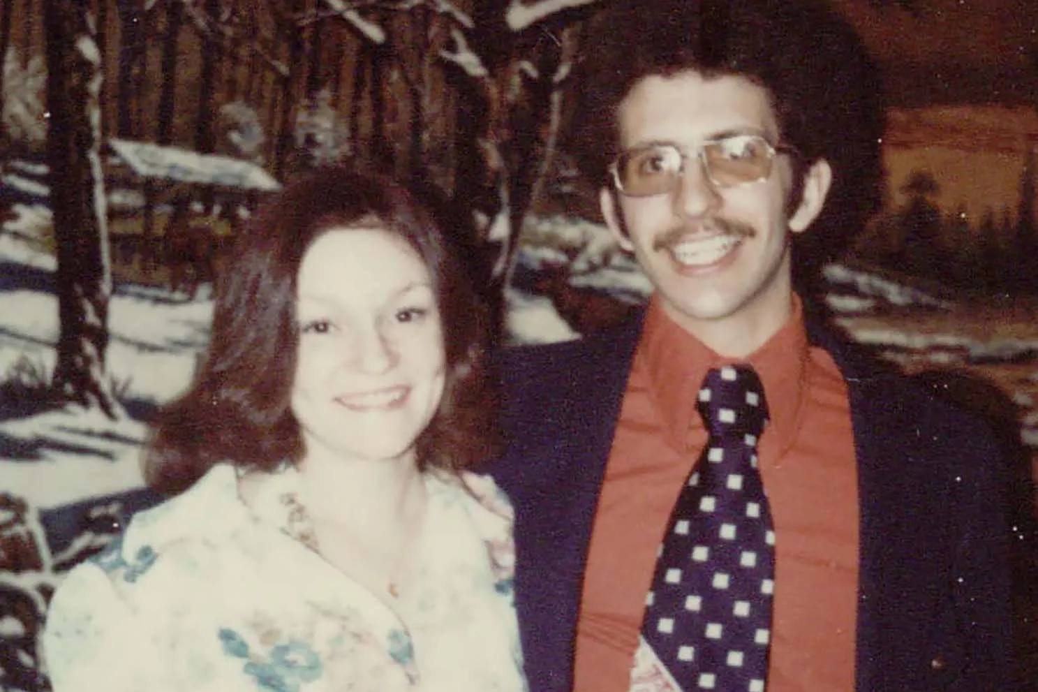 A couple poses for a photo smiling and dressed up. She wears a dress. He wears a tie with cubes, glasses and curly hair. Both are white a college-age.