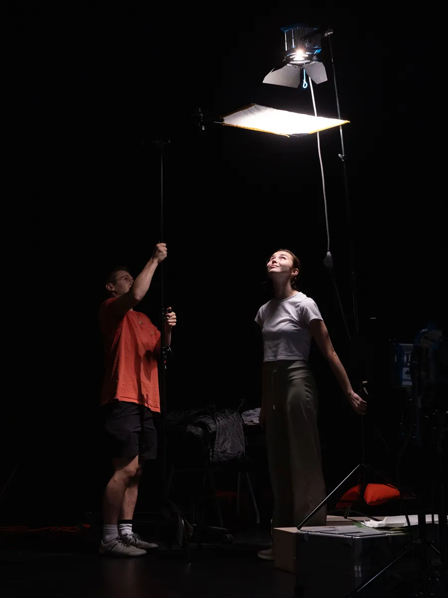 In a dark room, two students work under a light. The young man uses a long tool to hold a light diffuser under the light. The young woman stands looking up; the light on her face and body make her the most visible thing in the room.