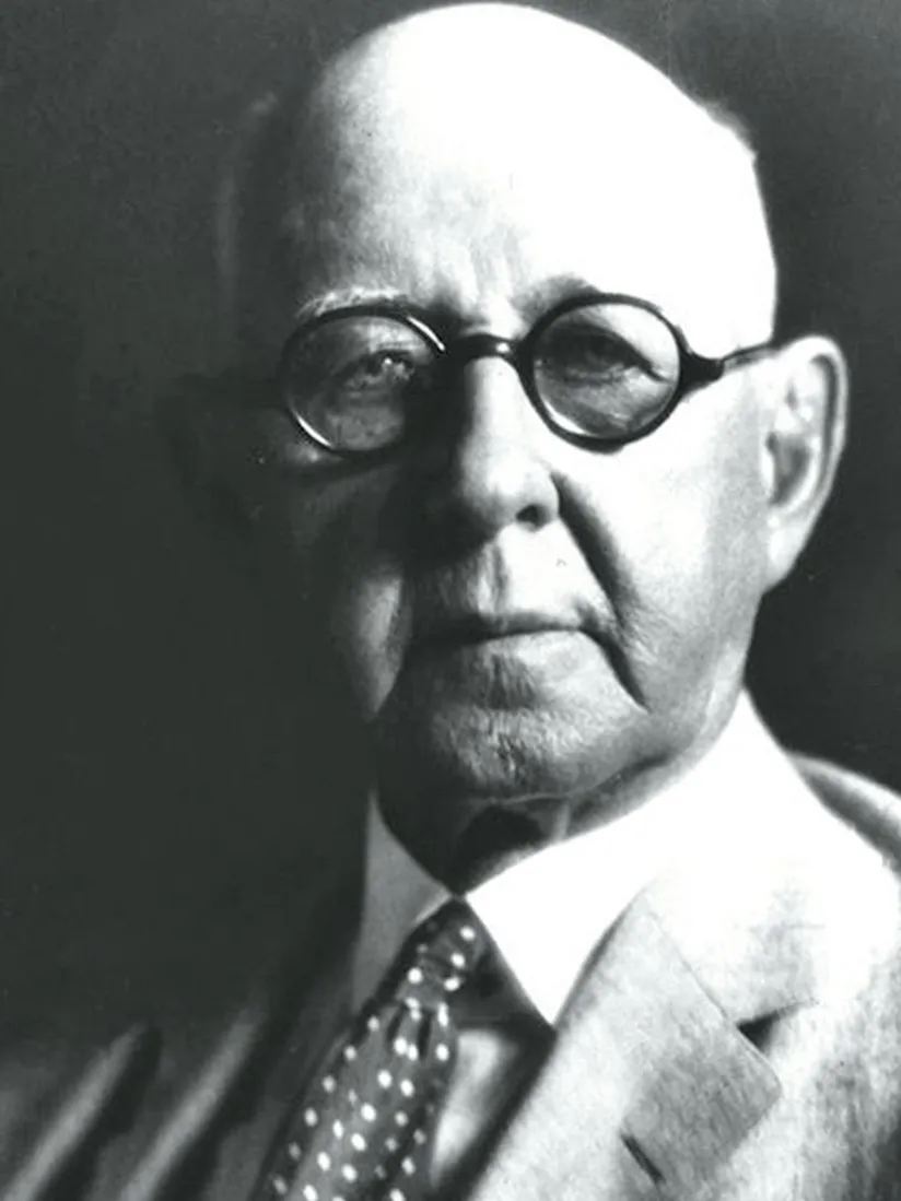 A distinguished looking man wears a suit, polka dot tie and round glasses. He is white and balding with a serious expression. This black and white photo of him is from the early 1900s.