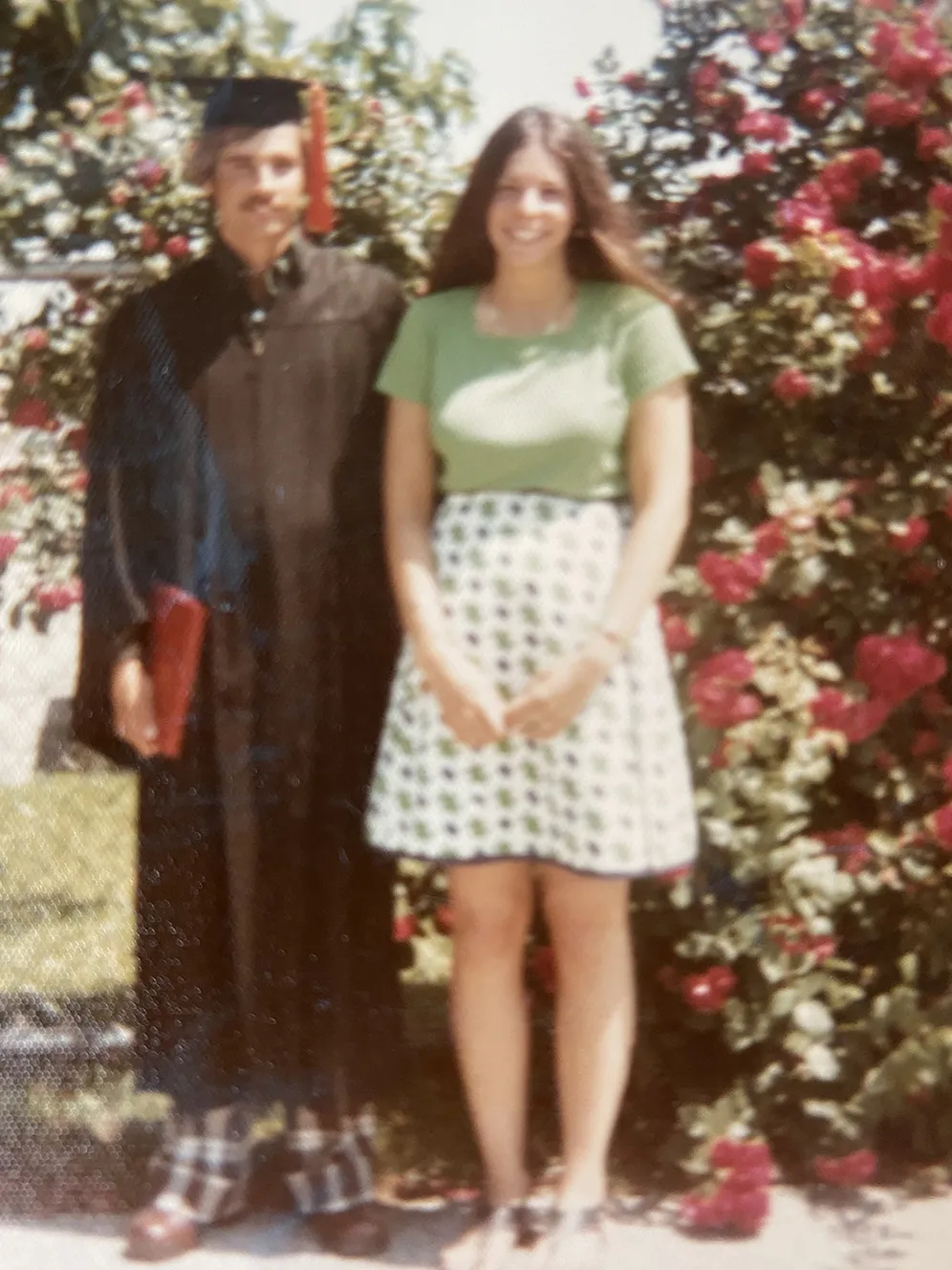 In a photo from the 1970s, a white man wearing a cap and gown stands next to his girlfriend. She is a young white woman with long brown hair and a pretty shirt and skirt.