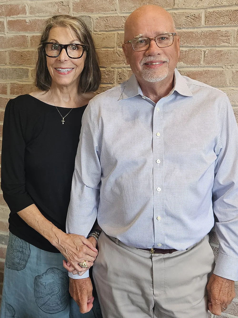 Now older, the same couple poses for a photo at a restaurant. They both now wear glasses, and have shorter hair. Hers is just past her chin; he's mostly bald. Both are happily smiling.