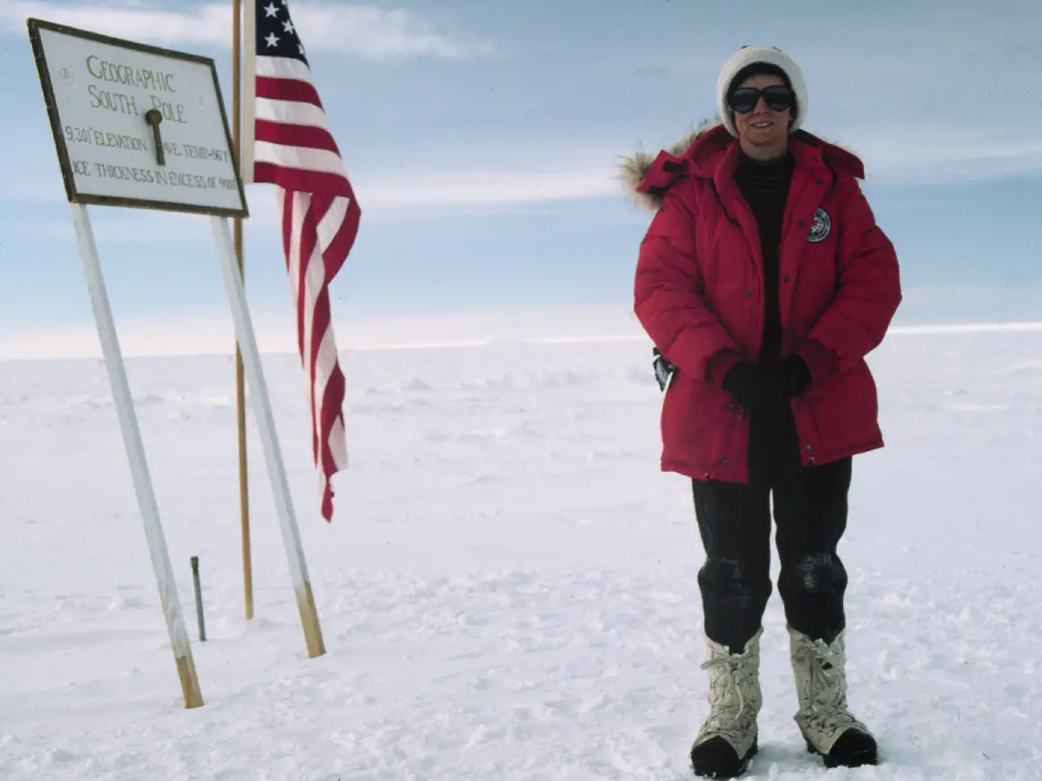 woman in red winter coat standing in snow at the south pole marker