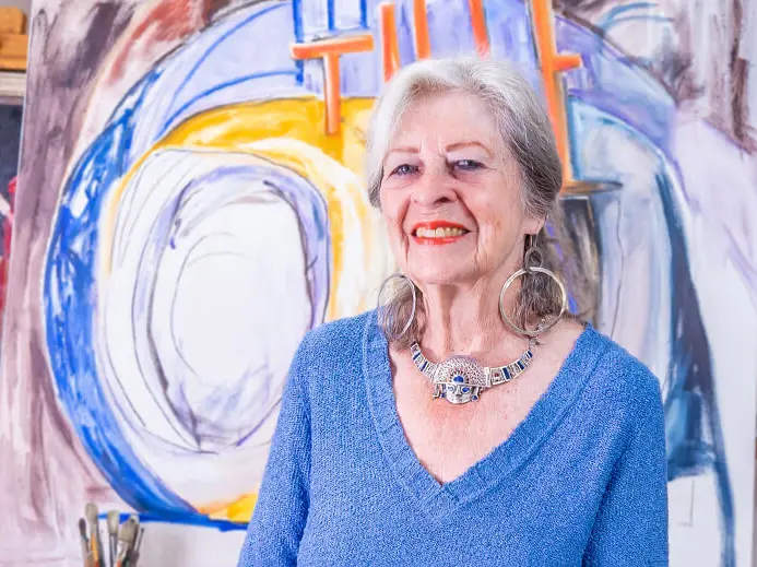 older woman in blue smiling while standing in front of artwork