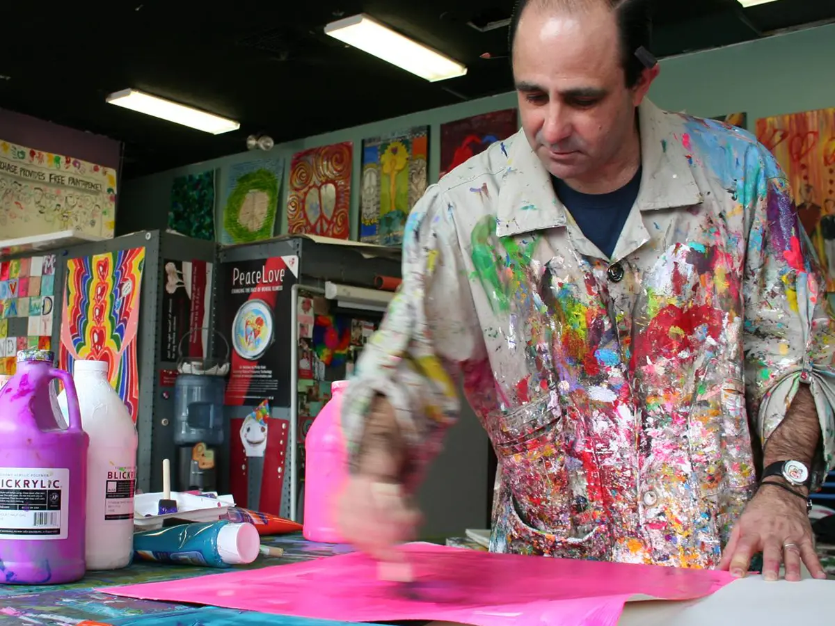 Wearing a paint-splattered smock, a man smiles slightly while painting in his colorful studio.