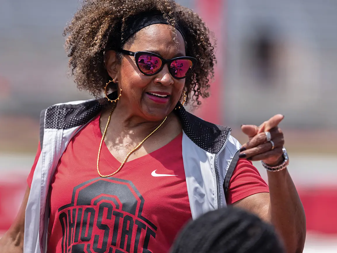 An older black woman wearing an Ohio State T-shirt, mirrored sunglasses, gold jewelry and a smile points while coaching outside.
