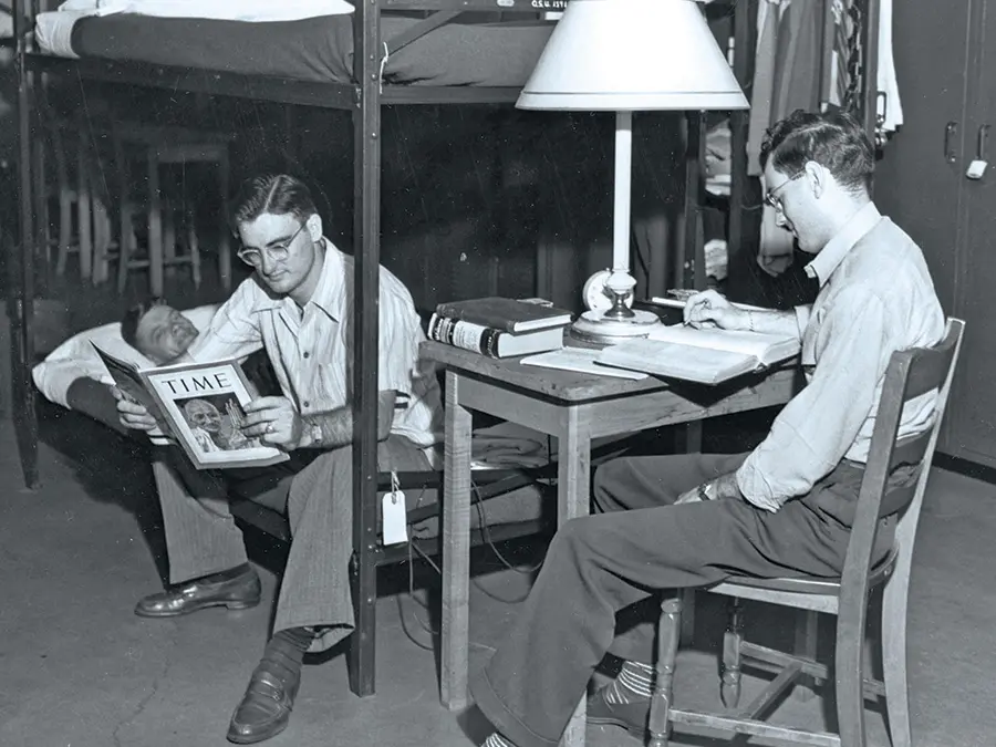 An old black and white photo shows bare-bones accommodations: A man studying sits at a small table. Another sits on a bottom bunk bed another man is lying in. A man in the background opens a locker where his clothes hang.