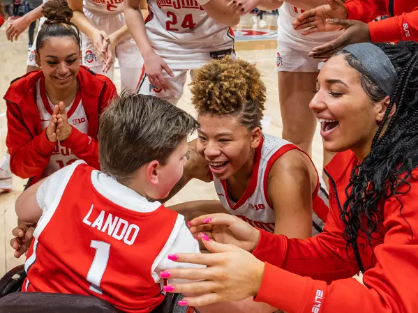 Ohio State women’s basketball players circle around a boy in a wheelchair, who is wearing a team and seen from behind. The women are smiling.