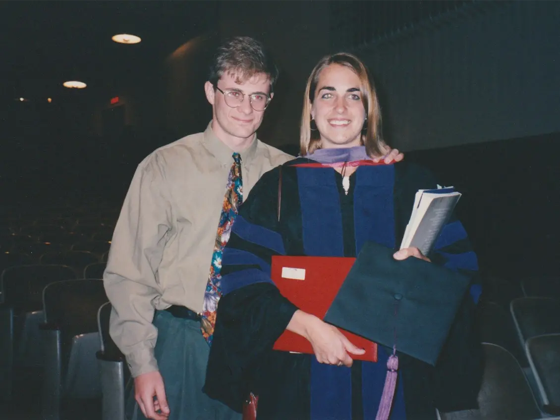 A young blond woman smiles proudly as she stands in her graduation gown holding her diploma and graduation cap. Her boyfriend, a young white man, stands behind her, also looking proud.
