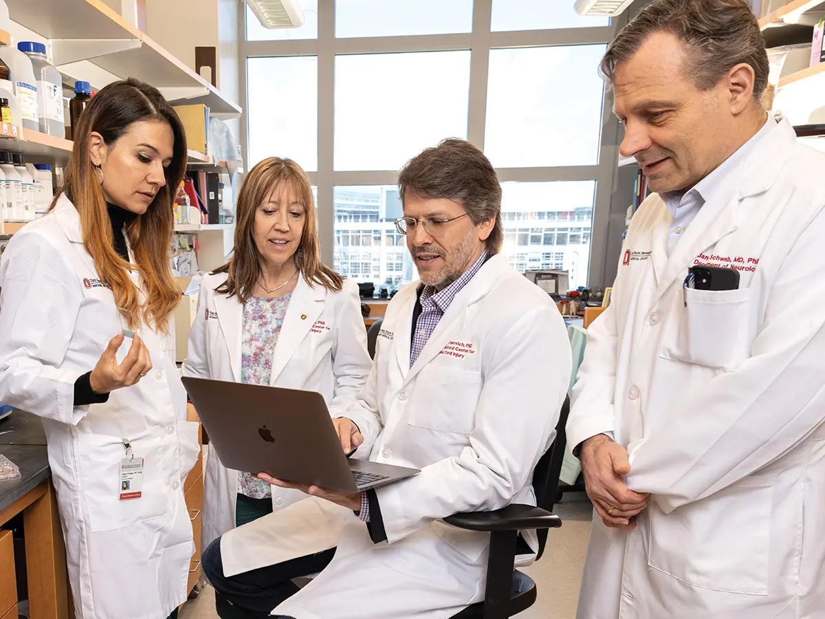 Four researchers wearing lab coats talk in a room filled with shelves of medications. Two are men and two are women.