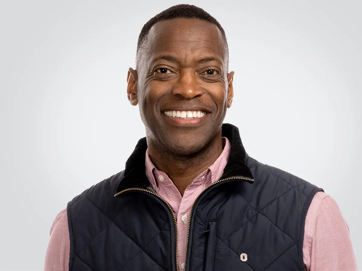 A black man with short hair, a wide smile and very symmetrical facial features looks happy yet relaxed while posing for the photo. His right hand is tucked into a pocket of his khakis, and he also wears a button-down shirt and zip-up vest.