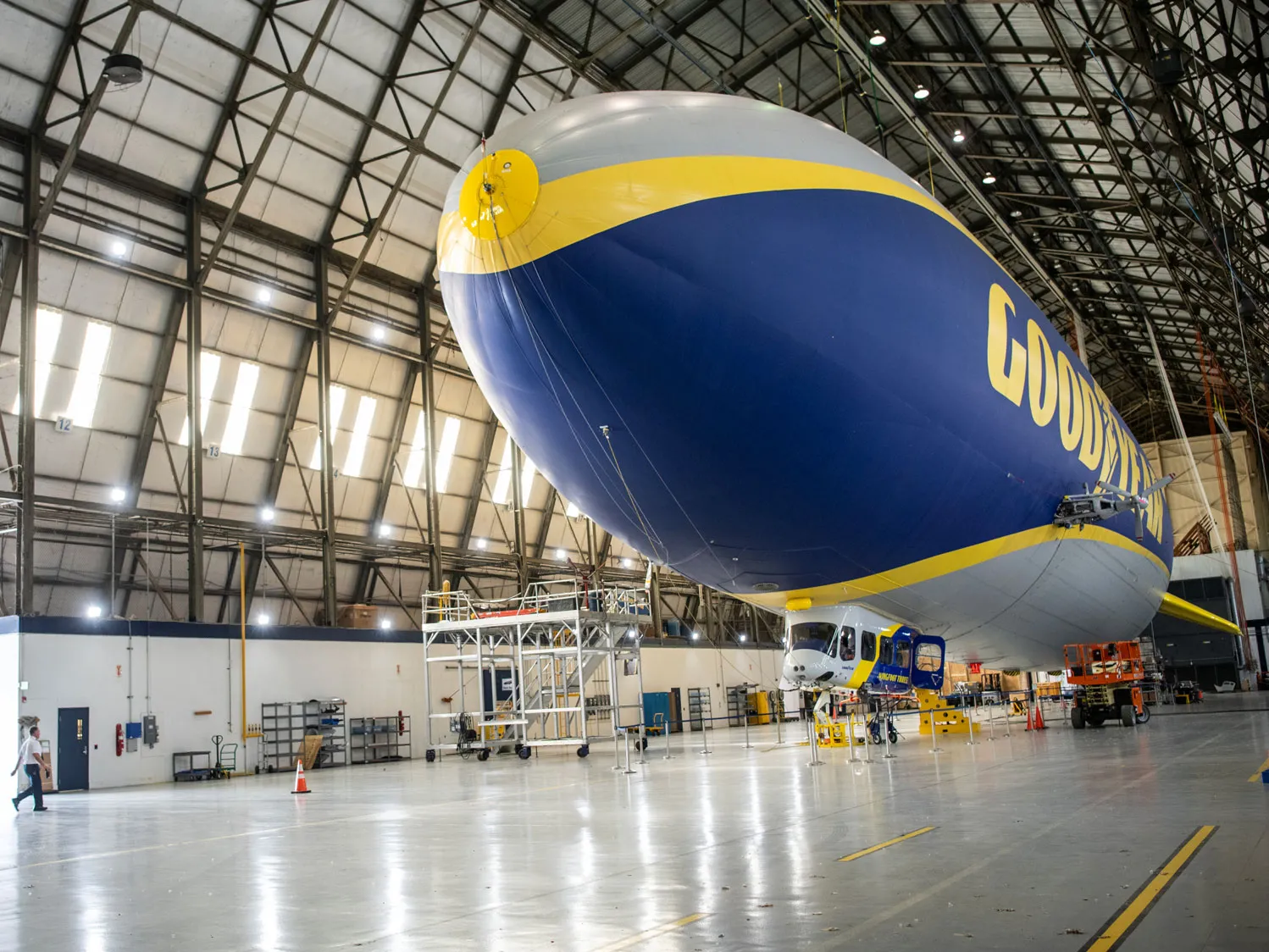 In a hangar with metal scaffolding supporting the 6-sided roof, a Goodyear blimp floats, looking a bit like a stamen inside a tulip. The floor is so clean, lights shine off it.