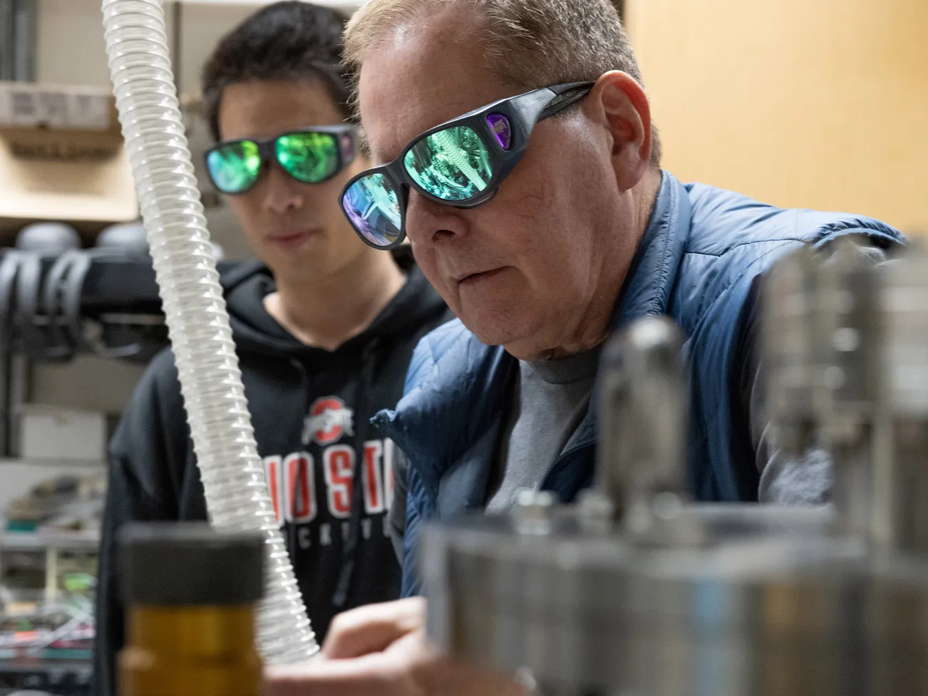 Both wearing safety goggles, Lou DiMauro and Yaguo Tang talk about a Quantum Trajectory Selector, which is the apparatus that lets them shoot lasers into gas to track electrons. Metal canisters and plastic tubes can be seen.