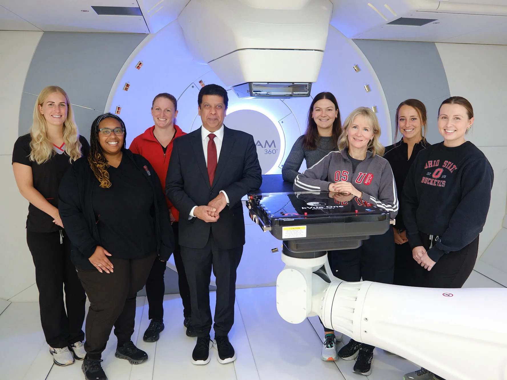 A team of people who work at Ohio State’s proton therapy center pose in the room where the treatment takes place. It is white-walled and appears spotless. They are eight smiling people—seven women—wearing scrubs or Ohio State sweatshirts. Dr. Arnab Chakravarti stands near the center in a suit.