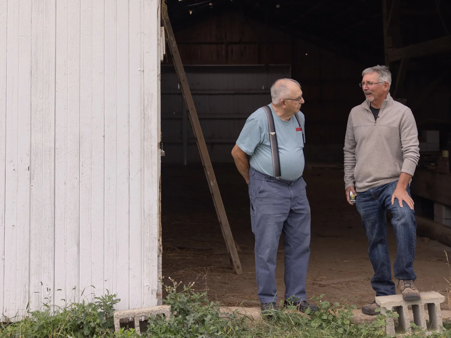 Two men stand chatting just inside opened barn doors. One is an older man wearing suspenders who owns the barn; the other is Doug Morgan who makes a living renovating barns and turning them into beautiful places for people to gather.