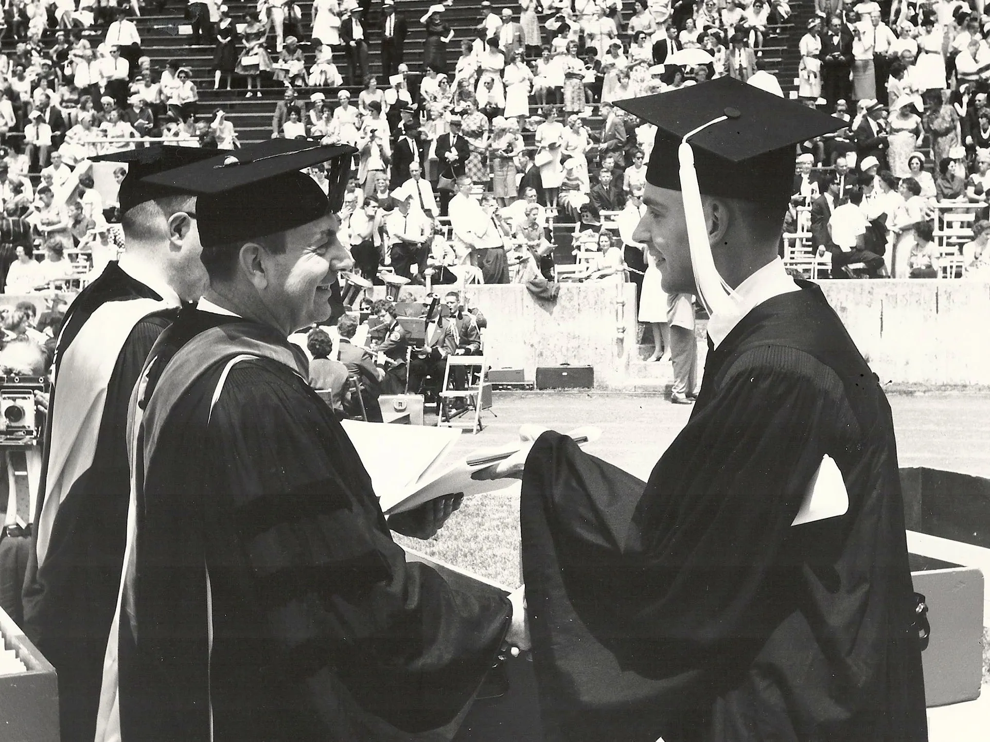 In a historical photo taken inside Ohio Stadium during a graduation commencement ceremony, a young man happily accepts his diploma from a pair of university leaders. All wear caps and gowns and smile. In the background, a crowd of spectators watches from the stands.