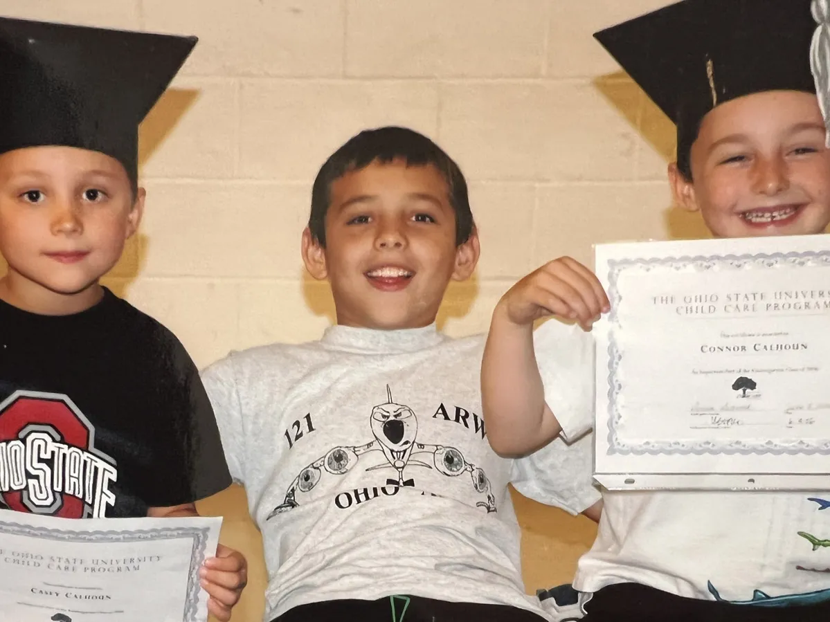 On a bench sit three boys. Two wear graduation caps and show off certificates, and the one between them has his arms wrapped around his brothers’ shoulders. He’s smiling, as is the boy on his left. The expression of the one on the right says he is less enthused, at least at the moment the photo was snapped.