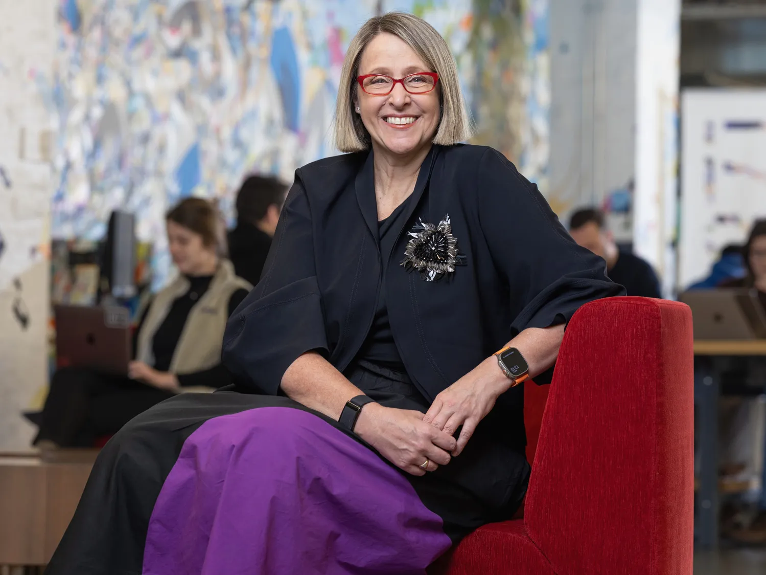 Betsy Ziegler sits on a modern upholstered chair in a space where many people in the background work or chat at small tables. A wall-size painting in the background adds to the lively feel of the scene, while Betsy poses with a genuine smile at the center of it. The middle-age white woman looks comfortable and stylish, with a chin-length haircut, stand-out glasses, modern-shaped clothes and a striking beaded broach that shines but also looks organically shaped. 