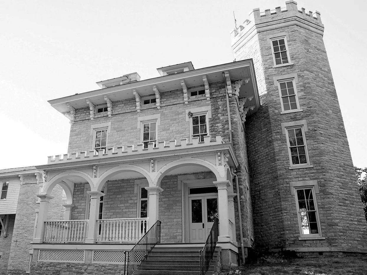 This old black and white photo shows cooke castle, a two-story stone building with fancy porch and a four-story tower extending on the right side, if you're a viewer facing the home.