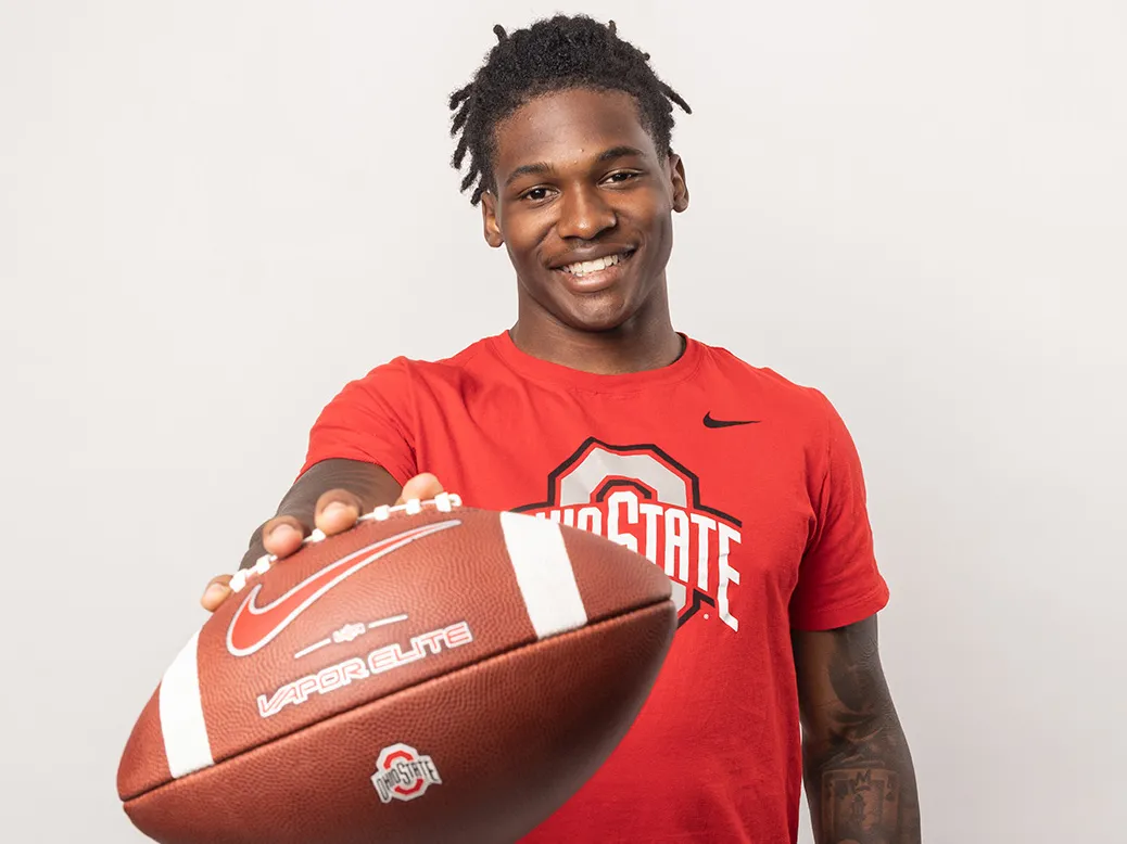 Ohio State football player Jeremiah Smith poses for a photo holding out a football. He's an attractive young black man, smiling with his hair pulled up in a ponytail and tattoos on his arms.