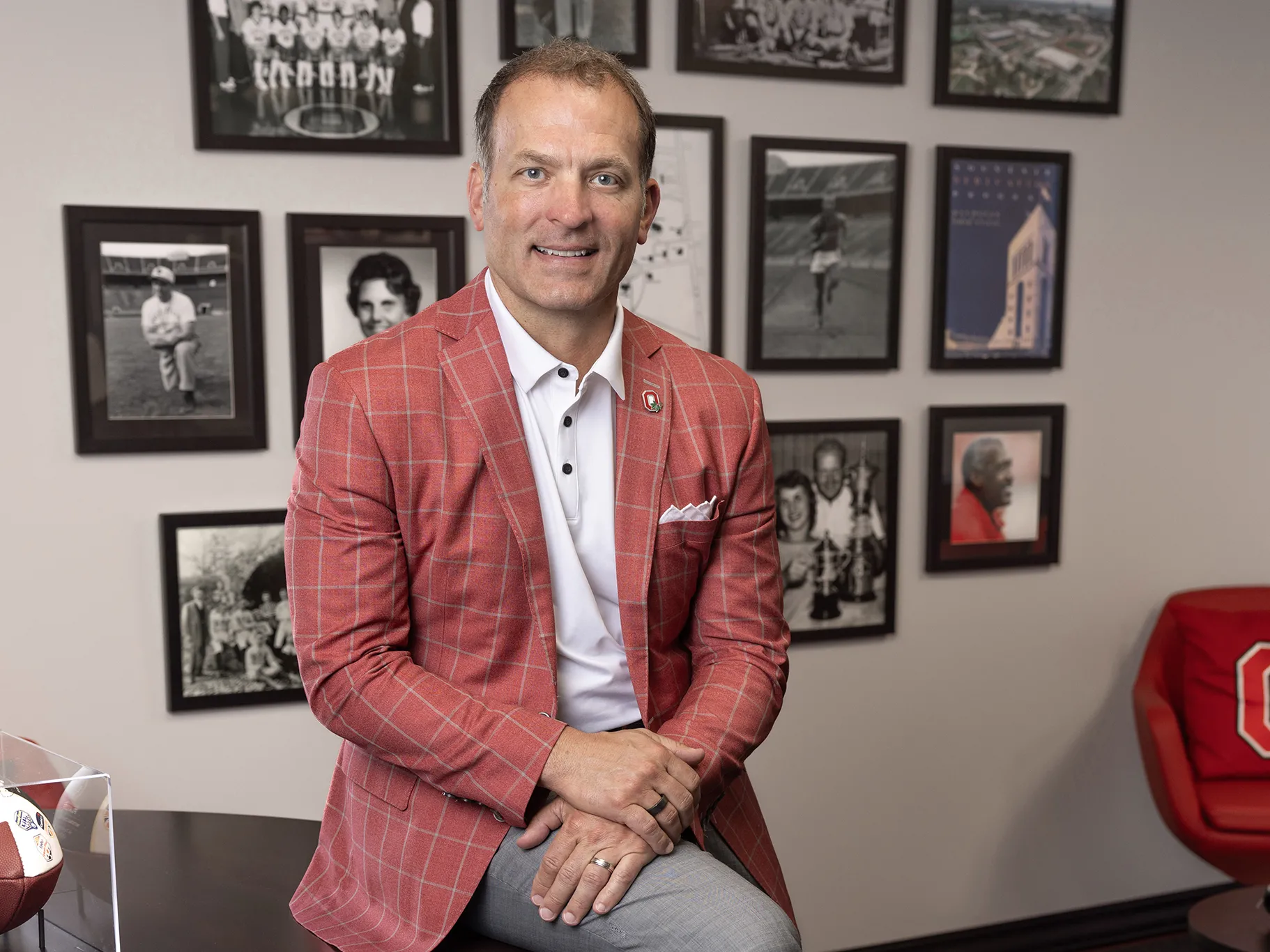 Ohio State Athletics Director Ross Bjork smiles as he poses for a photo in his office. On the wall behind his are photos of great people and moments in Ohio State sports history.