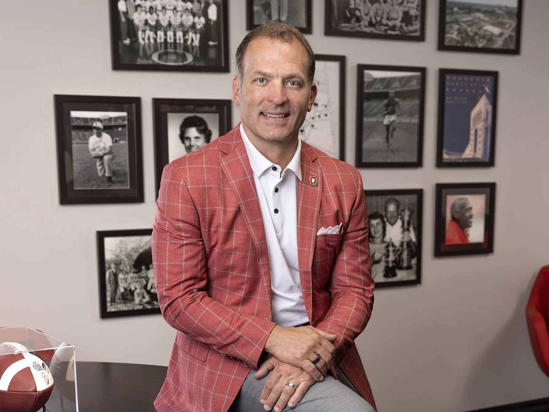 Ohio State Athletics Director Ross Bjork smiles as he poses for a photo in his office. On the wall behind his are photos of great people and moments in Ohio State sports history.