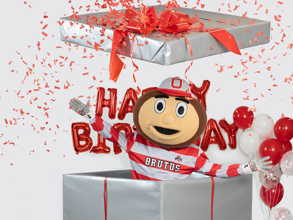 In this Ohio State photo, Brutus Buckeye jumps out of a giant wrapped birthday present. Balloons in Ohio State's colors, as well as shiny mylar balloons that say happy birthday, are around him and confetti flies everywhere. Brutus is the university's mascot--a giant head shaped like a buckeye tree nut with a big grin.