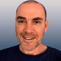 A portrait of Steve Wartenberg shows a white man with brown eyes and beard stubble smiling, as if he's hearing something that will make him laugh momentarily.