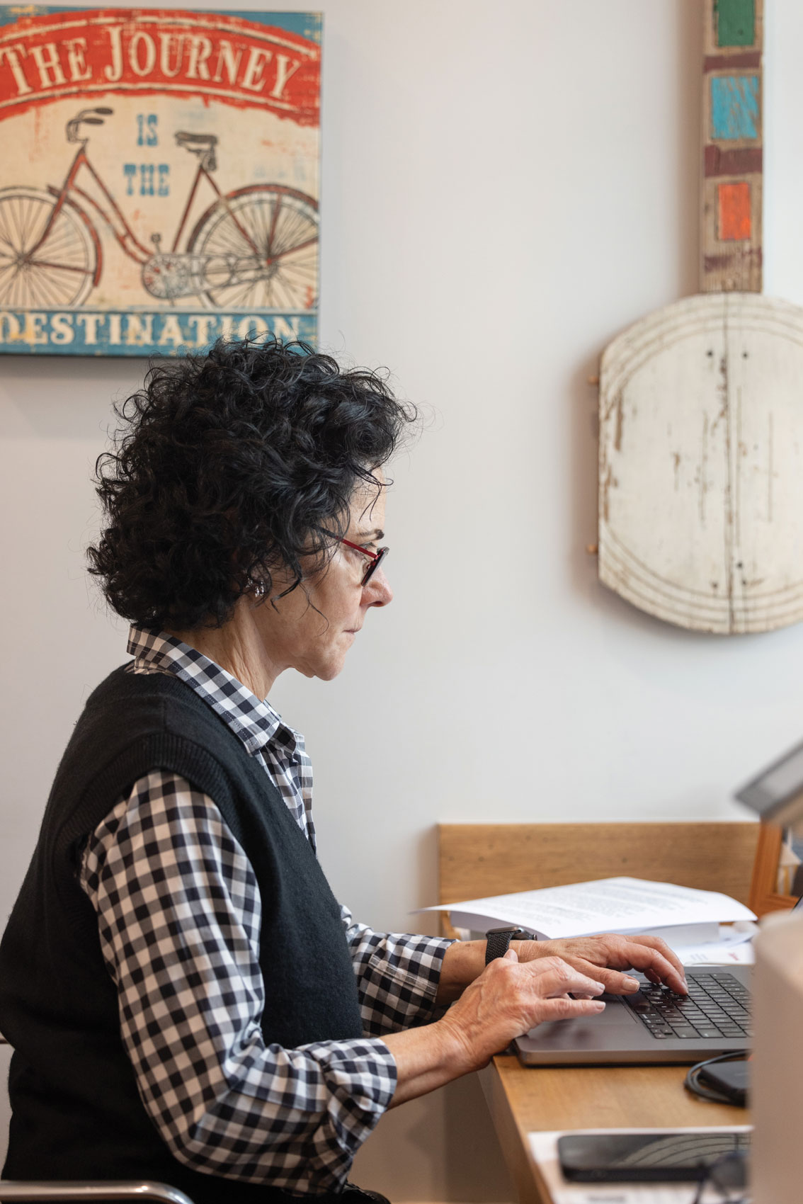 Wearing a plaid shirt and sweater vest, Linda sits at her desk at home, working on a laptop.