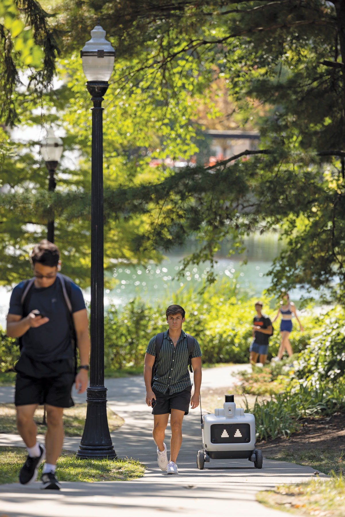 AN a pretty walk from Mirror Lake a food-delivery robot rolls up the hill alongside a young man wearing a backpack.