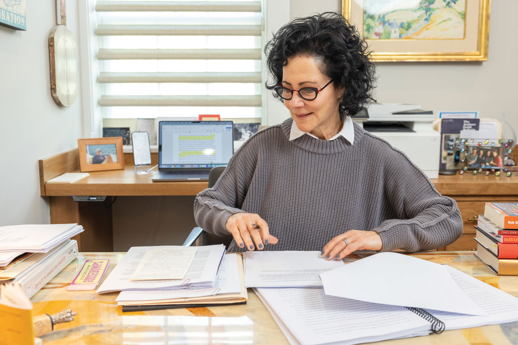 Wearing stylish reading glasses and a gray pullover sweater, Linda works in her home office. A wire bound copy of her manuscript and other printed-out stacks of paper are before her.