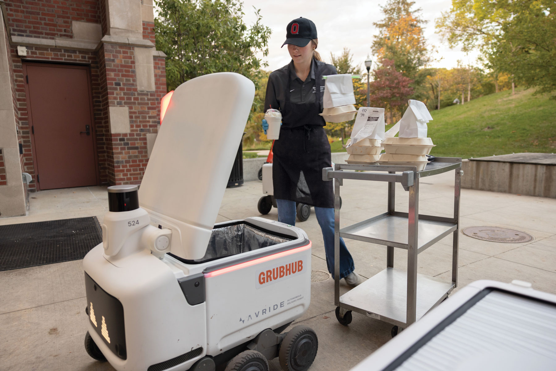 A restaurant worker wearing an aprob and hat moves food from a metal cart into an open robot. It's back hatch is opened like a clamshell.
