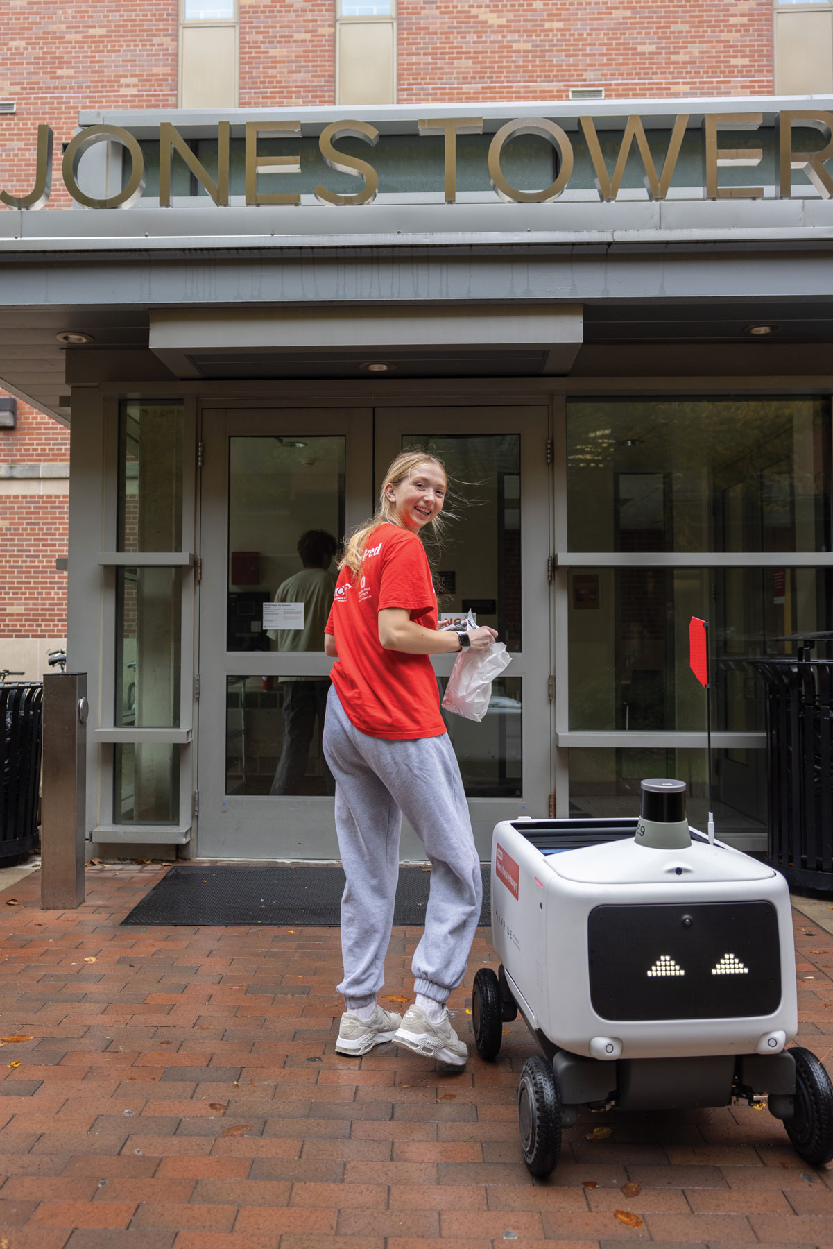 A young woman looks over her shoulder and smiles for the photographer as she takes her food from the robot to the door of her residence hall.
