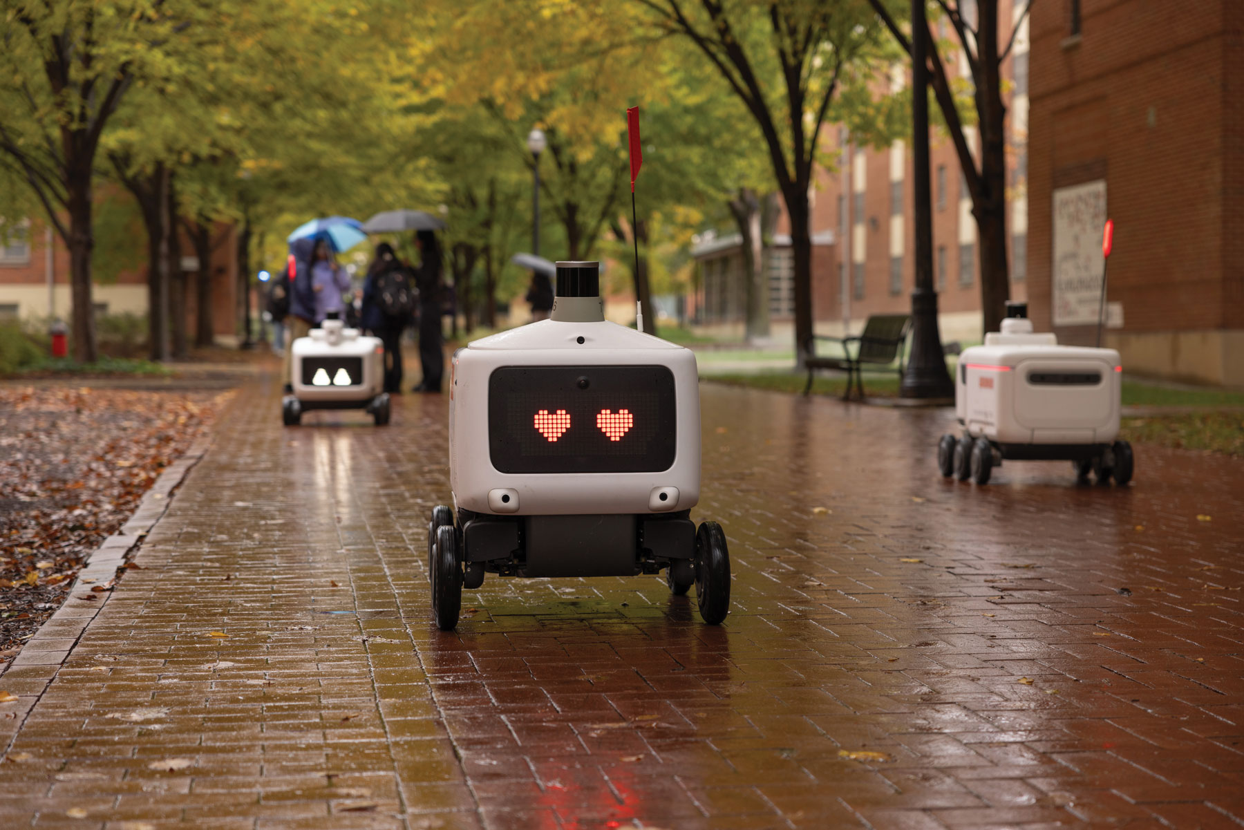On a wet brick sidewalk, a robot has heart eyes and rolls merrily on its way. They things closest to it are other robots; students in the distance carry umbrellas.