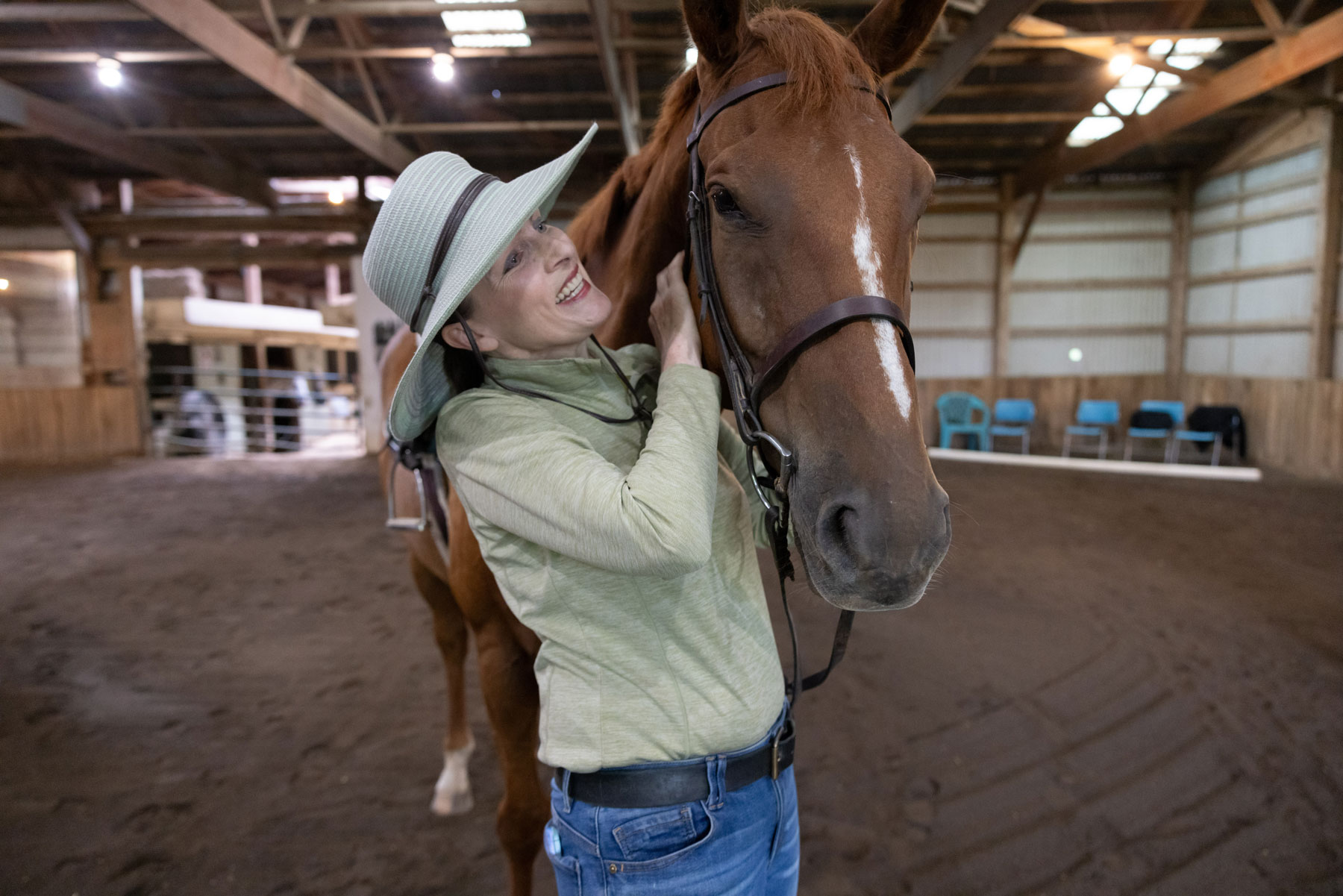 Lloyd smiles as she wears a wide-brimmed hat and stands close to a horse, holding its head gently while standing inside an indoor riding arena.