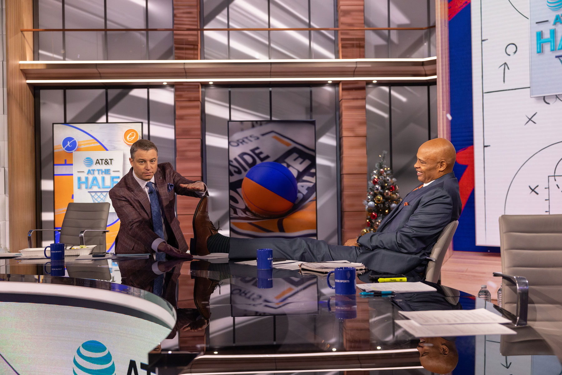 Between segments, Kellogg leans back in his broadcast chair, stretching his long legs onto the broadcast desk. Zuckers holds his hands up, measuring how long Kellogg’s foot is. They’re both chuckling.