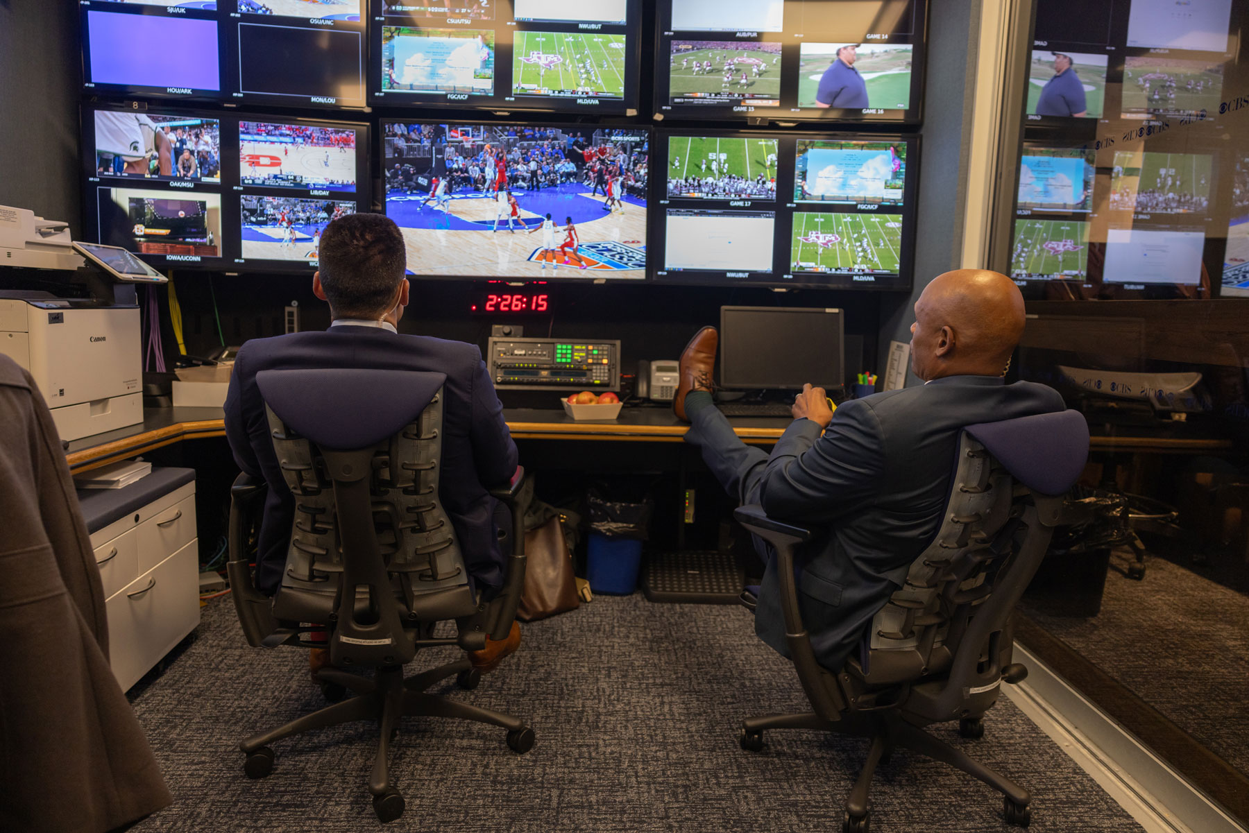 Seen from behind, Kellogg and Szczerbiak sit in desk chairs inside a control room, facing a wall of screens showing basketball games. Szczerbiak sits upright, while Kellogg leans back with one leg propped on the desk.