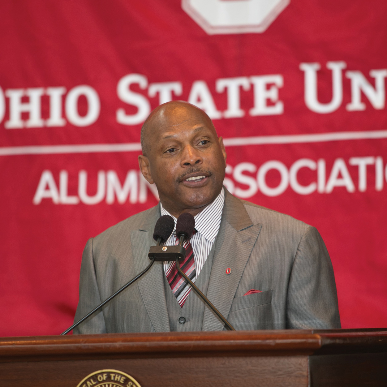 In a three-piece suit, Griffin stands behind a lectern with two microphones. Behind him is a giant banner that says “Ohio State University Alumni Association.” He’s smiling while he listens to someone off camera talking or asking a question.