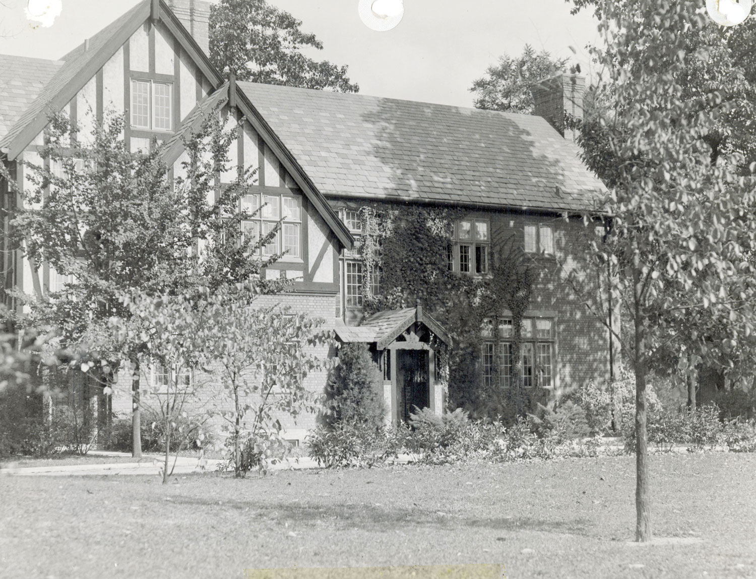 A large house with steep gabled roofs and timber-accented stucco exterior is shown from the front, partly covered by leafy trees and climbing plants. The home has multiple windows and a small covered entryway. 