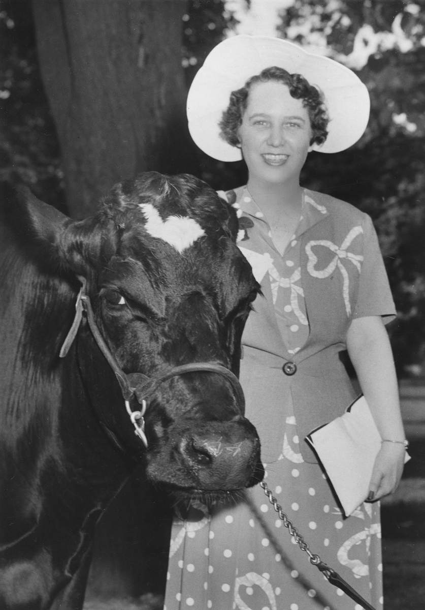 A woman dressed in a dress and wide brimmed hat smiles as she poses with a cow in this black and white photo from 1942.