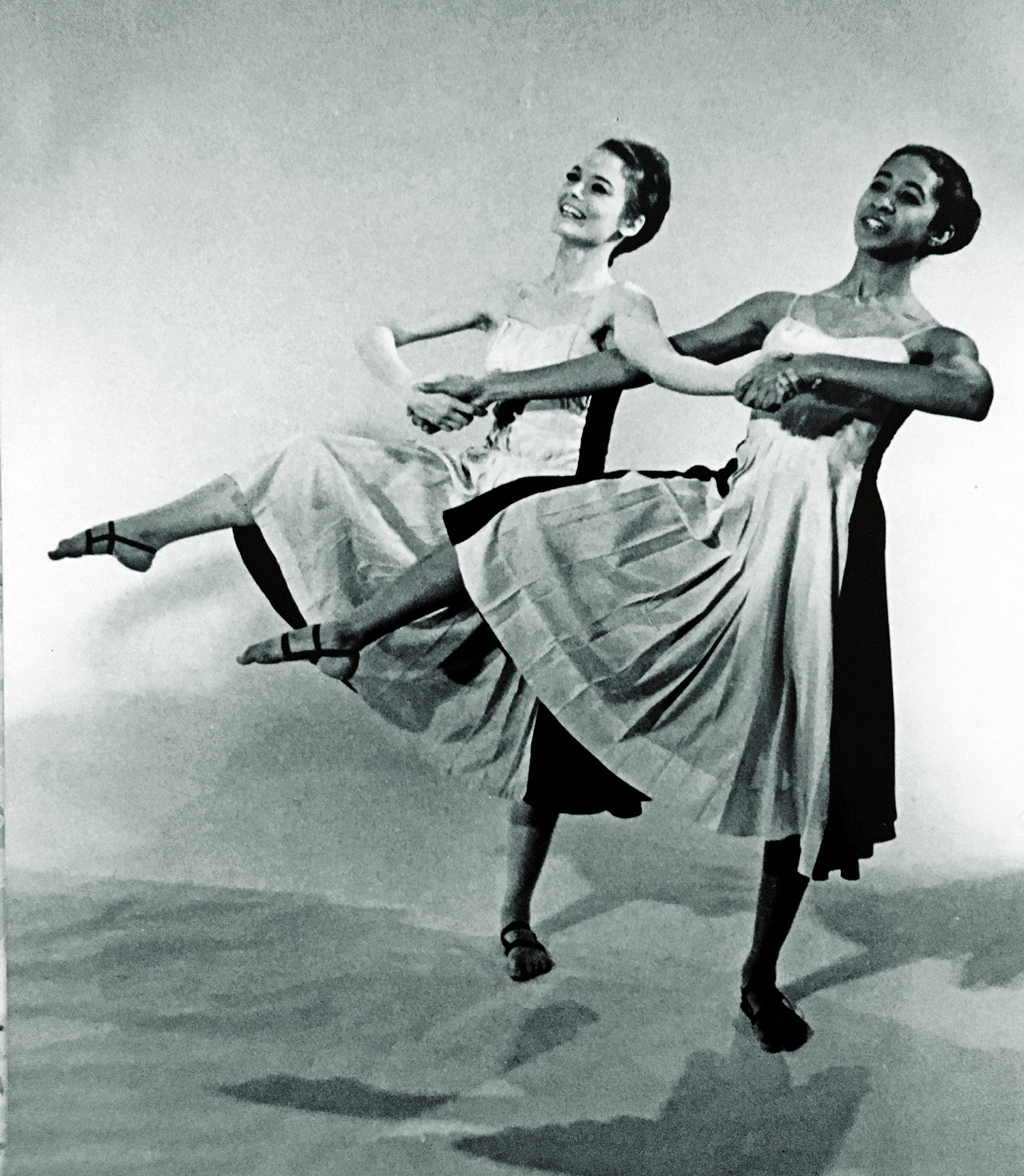 In a black and white image from the 1960s, two women, one white and one black, link hands and arms as they dance together. Both have pretty smiles, their right leg lifted and bent, toes pointed, and pleated-skirt dresses. Their bodies are moving exactly the same way and are tilted in exactly the same angle.