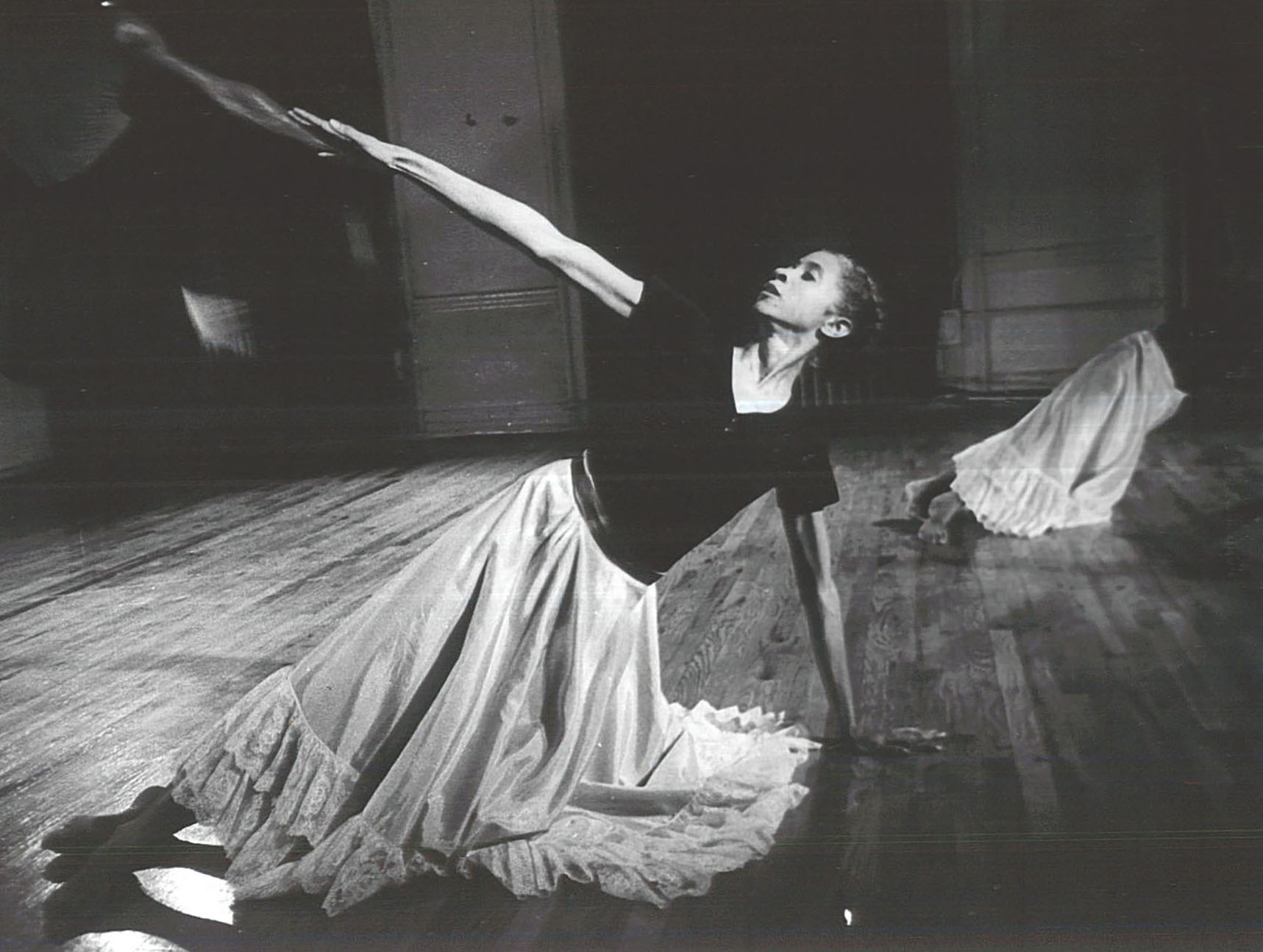 In a dance studio, Diane plants one arm on the ground and leans her body into it, stretching out her other arm as far as she can. Her chin is lifted as she looks toward her outstretched hand; her body is a straight line down to her crossed feet. Her shin-length satin dance skirt, with bottom ruffles, pools on the ground. She appears both strong and graceful.