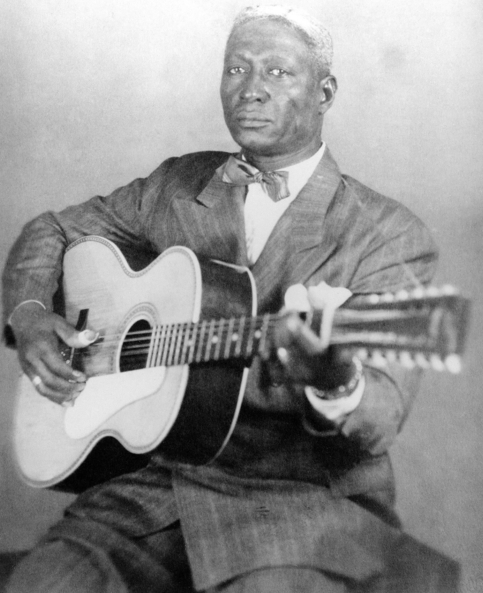 A black man with white hair holds a guitar and wears a pin-striped suit and bow tie. His expression is serious--almost sad in the way his eyes look directly into the camera.