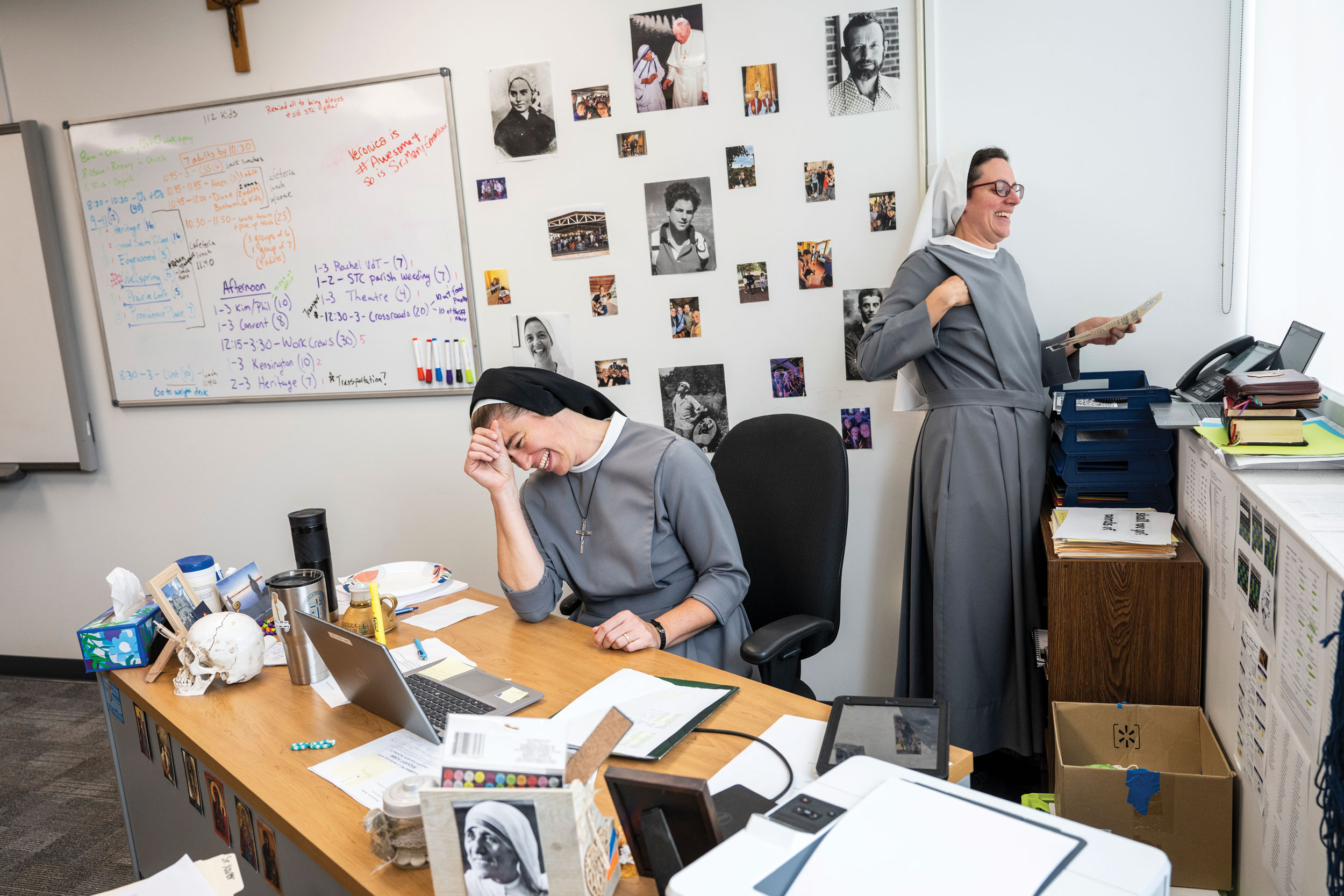 Two sisters laugh in a school office as they carry out administrative tasks. On the wall behind them are photos of people they admire, including popes and saints.