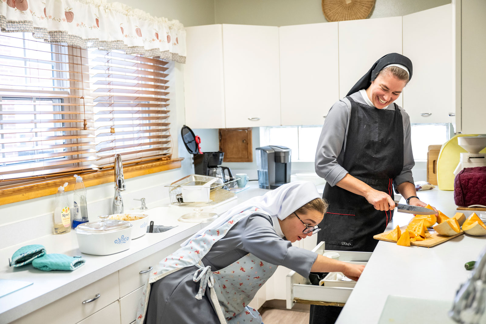 Two sisters cut up squash in a kitchen. Sister Xavier laughs as the other sister looks for something in a drawer.