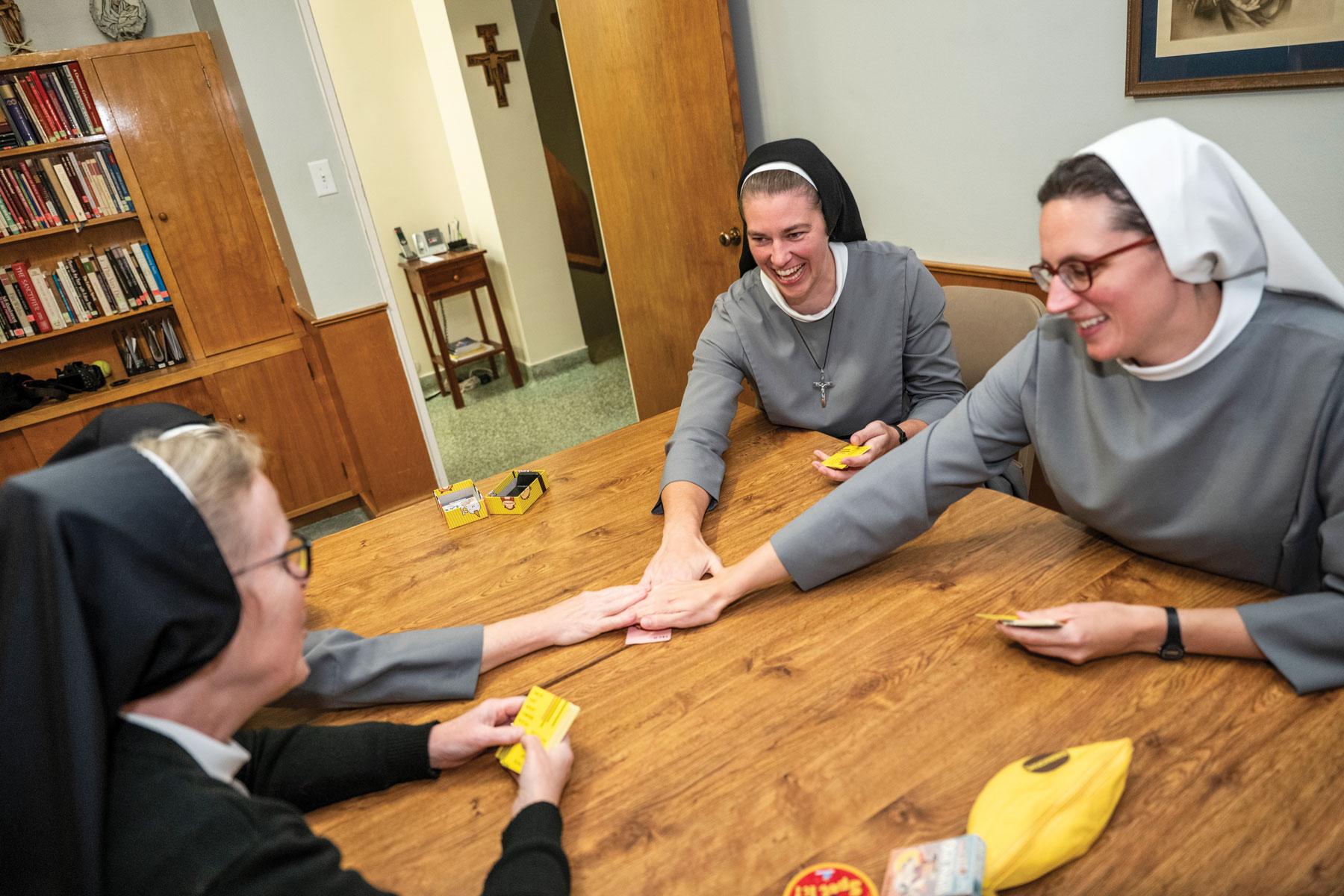 Sisters play a card game in their home. The decor is plain and serviceable, but the sisters seem to be having a good time sitting around the table. 