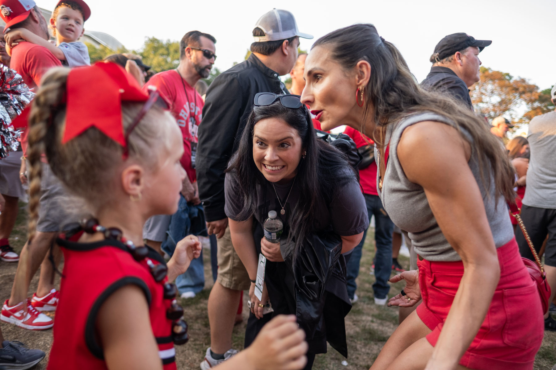 Melissa grins as she bends forward to talk to a young girl at face level. The girls is wearing a Buckeyes cheer costume and another woman talks with them while a crowd swirls around them.