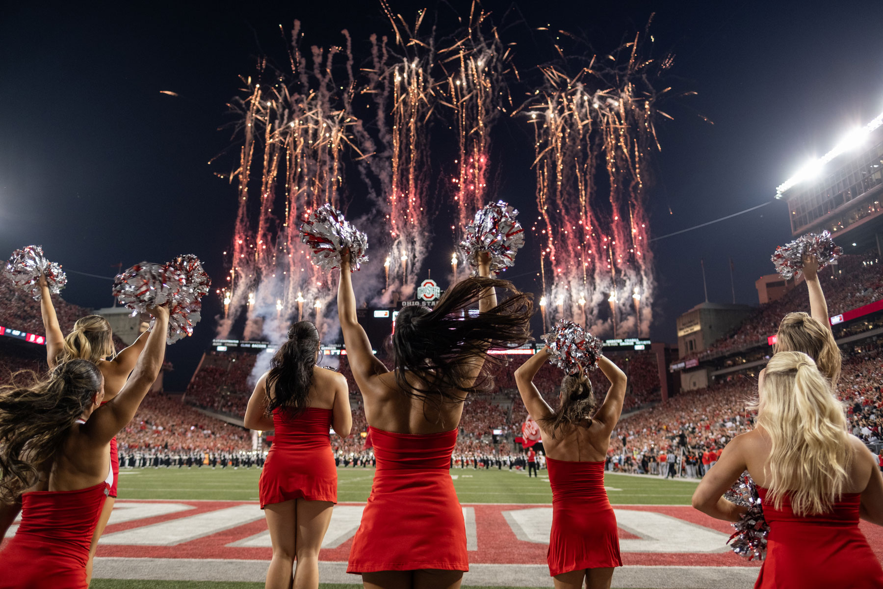 Seen from behind, the dance team jumps and waves their pompoms as fireworks shoot up across the football field. It's dark out but the stadium is lit up.