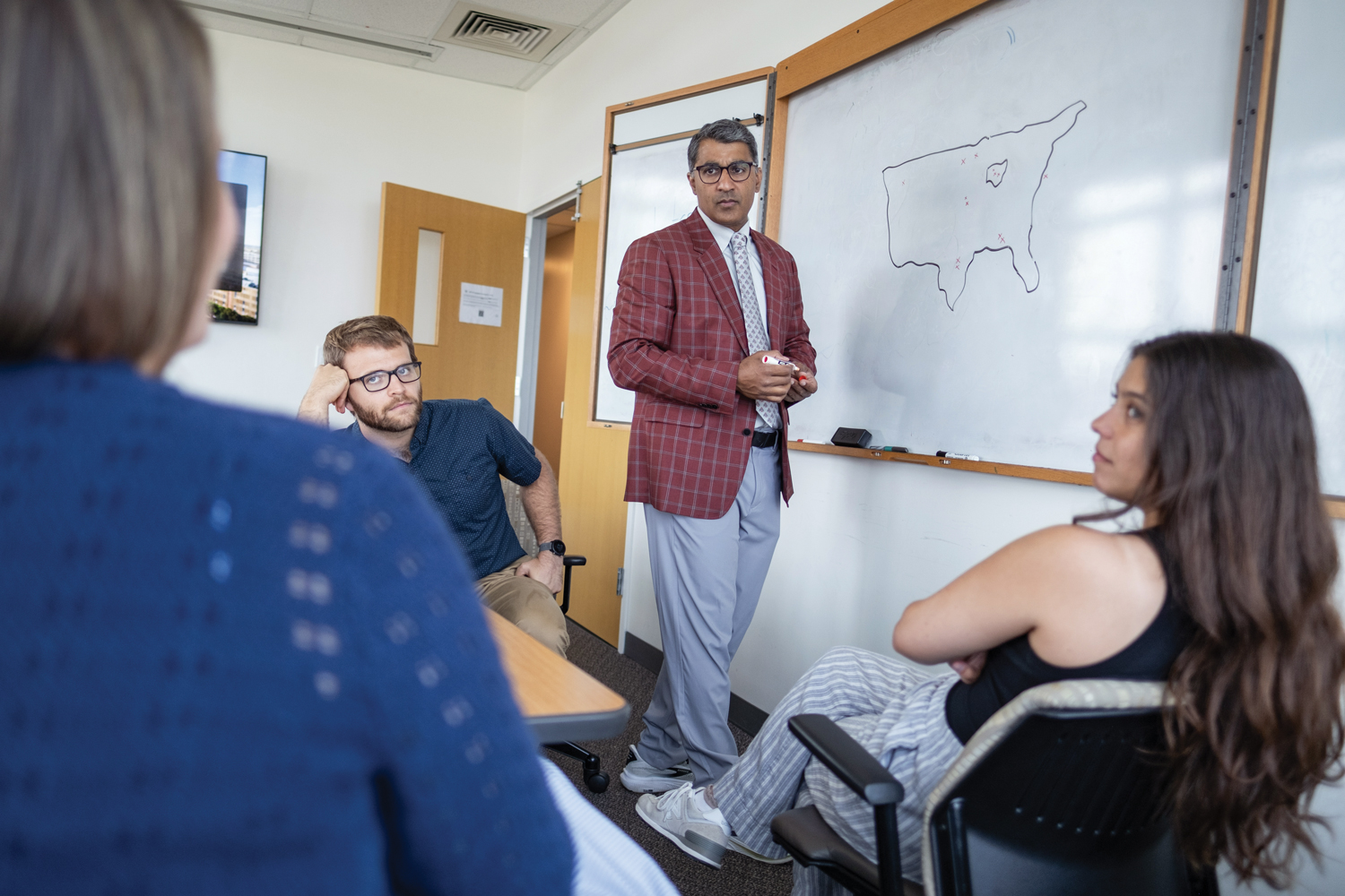 Roychowdhury stands at a white board holding a marker while he listens to a team member. Two more also look at her as she talks.